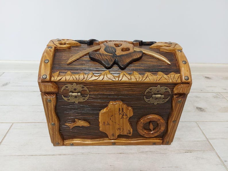 May include: A wooden treasure chest with a dark brown finish and carved details. The chest features a map, compass, and fish design on the front. The lid has a carved skull and crossed swords. The chest is 12 inches long, 8 inches wide, and 8 inches tall.