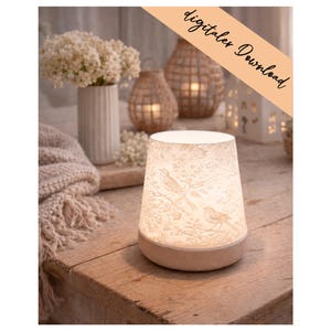 Pode incluir: Uma lâmpada decorativa com base bege e abajur branco estampado com pássaros e folhagens. A lâmpada está sobre uma mesa de madeira, com um vaso de flores brancas e outros objetos decorativos no fundo. O texto "digitaler Download" é visível.