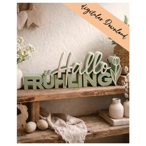 Puede incluir: Un letrero de madera que dice "Hallo Frühling" en blanco y verde. La palabra "Hallo" está en cursiva, y "Frühling" en letras de imprenta. El letrero está decorado con un tulipán y se encuentra en una estantería de madera con otros objetos decorativos.