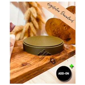 Puede incluir: Un soporte de exhibición de producto circular verde oliva con un diseño de montaña en la parte delantera. El soporte está sobre una tabla de cortar de madera. La imagen tiene el texto "digitaler Download" y "ADD-ON".