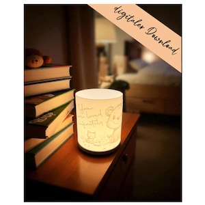 May include: A cylindrical, illuminated night light with a soft glow, featuring an etched design of a bear, a kitten, and the words "You are loved infinitely." The lamp sits on a wooden nightstand next to a stack of books.