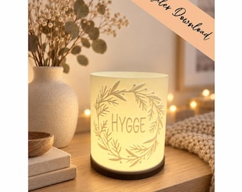 May include: A cylindrical white lamp with a dark base featuring a cutout wreath design and the word HYGGE in the centre. The lamp emits a warm glow and includes a diagonal banner in the upper right corner with the text digitaler Download.