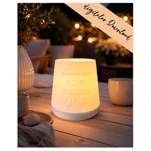 Op de afbeelding: Een oplichtende witte cilindervormige lamp met de tekst "Sommernächte duften immer ein bisschen nach Glück" staat op een houten tafel. De lamp geeft een warm licht. Andere items op de tafel zijn een keramische mok en een decoratief dienblad.