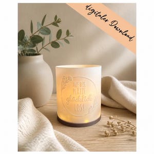 Puede incluir: Un portavelas cilíndrico de vidrio esmerilado con base oscura, iluminado desde el interior. El frente presenta un marco circular en relieve con el texto en alemán "AUF DIE PLÄTZE glücklich LOS!" y detalles florales. Una banda color melocotón dice "digitaler Download".