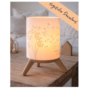 May include: A cylindrical lamp with a dandelion design, resting on three wooden legs. The lamp emits a warm glow, and the text "digitaler Download" is visible. The lamp is on a wooden table.