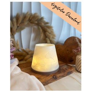 Puede incluir: Un portavelas de cerámica blanca con base marrón y diseño de hojas de ginkgo en relieve. El portavelas está encendido, emitiendo un brillo cálido. Las palabras "digitaler Download" están escritas en una pancarta diagonal color melocotón.