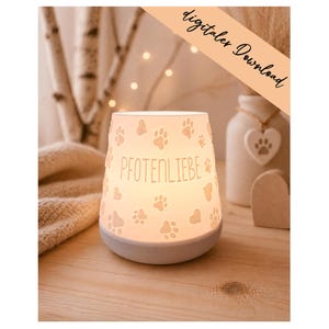 Op de afbeelding: Een verlichte witte keramische nachtlamp met pootafdrukken en het woord "PFOTENLIEBE". De nachtlamp staat op een houten oppervlak, met een zachte deken en decoratieve items op de achtergrond.