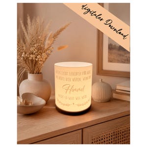 Puede incluir: Lámpara cilíndrica iluminada, con base negra y pantalla blanca con texto en alemán, incluyendo la palabra "Himmel" (Cielo). Se encuentra sobre una superficie de madera, con jarrones decorativos y plantas secas en el fondo.