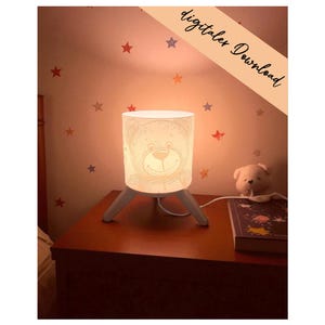 May include: A white cylindrical table lamp with a line-art illustration of a teddy bear face on the shade, supported by three light-colored wooden legs. The lamp emits a warm glow and features the text digitaler Download in the corner.