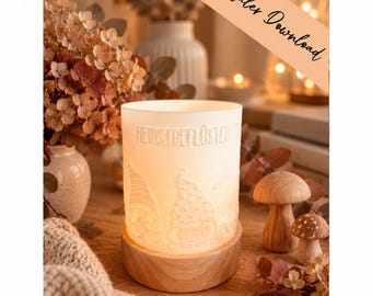 May include: A cylindrical white frosted glass candle holder featuring embossed gnome figures and the text HERBSTGEFLÜSTER, set on a circular light wood base. The glass is illuminated from within, casting a soft glow on the surrounding surface.