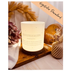 Può includere: Un portacandele cilindrico in ceramica bianca con la scritta "STOFFSTREICHLERIN" e "digitaler Download". Il portacandele è posto su una superficie di legno, con una base in metallo marrone scuro. Fiori secchi e fogliame sullo sfondo.