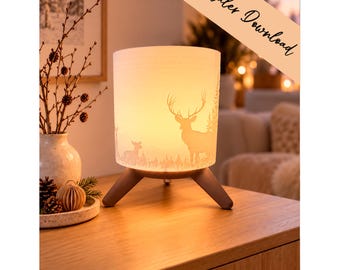 May include: A cylindrical table lamp with a warm glowing shade featuring a silhouette of a deer and forest landscape. The lamp has three dark brown wooden legs and includes a diagonal banner with the text digitaler Download.