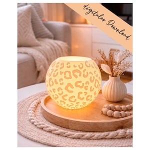 May include: A spherical lamp with a white leopard print pattern that emits a warm glow. The lamp sits on a round wooden tray alongside a small white ribbed vase containing dried flowers and a string of light-coloured wooden beads. Text reads digitaler Download.