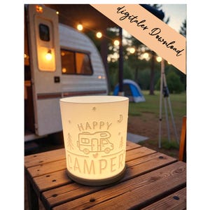 Könnte beinhalten: Eine leuchtende, weiße zylindrische Lampe mit den Worten "HAPPY CAMPER" und einer Wohnmobil-Illustration. Die Lampe steht auf einem Holztisch, im Hintergrund ein Wohnwagen und Lichterketten. Der Text "digitaler Download" befindet sich oben rechts.