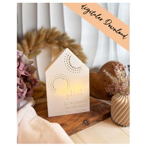 Può includere: Un portacandele bianco a forma di casa con ritagli decorativi e la parola "glück" illuminata. Il testo "IST KOSTENLOS ABER DENNOCH UNBEZAHLBAR" è sotto. L'immagine include anche fiori secchi e un cuore di legno.