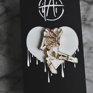May include: Gold-tone enamel pin featuring a white skeleton tied up with red rope. The pin is set against a white heart-shaped background with dripping paint.