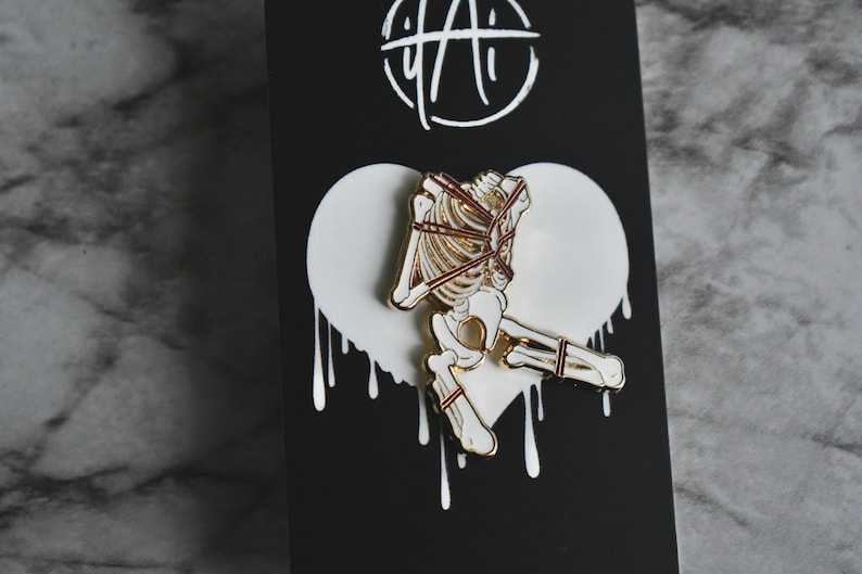 May include: Gold-tone enamel pin featuring a white skeleton tied up with red rope. The pin is set against a white heart-shaped background with dripping paint.