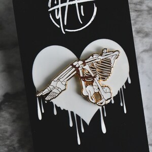 May include: A white enamel pin with a gold outline depicting a skeleton tied up in a heart shape. The heart is dripping with white paint.