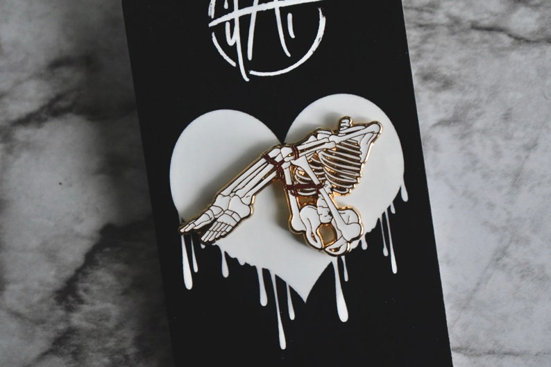 May include: A white enamel pin with a gold outline depicting a skeleton tied up in a heart shape. The heart is dripping with white paint.