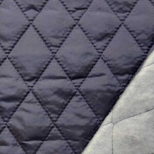 Op de afbeelding: Close-up van twee gewatteerde stoffen. De ene is een diep marineblauw met een glanzende afwerking, gestikt in een ruitpatroon. De andere is een lichtgrijze, matte stof met dezelfde ruitvormige quilting. De stoffen zijn waarschijnlijk voor naaiprojecten.