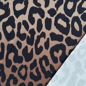 Può includere: Primo piano di un tessuto con stampa leopardata. Il tessuto presenta uno sfondo marrone con macchie nere. Una porzione del tessuto è ripiegata, rivelando un rovescio azzurro chiaro con un motivo leopardato tenue.