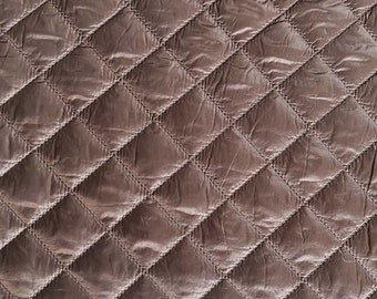 Puede incluir: Primer plano de una tela acolchada con un patrón de diamantes. La tela es de color marrón apagado, con costuras visibles que delinean cada diamante. La textura parece suave y ligeramente brillante, lo que sugiere un material acolchado.