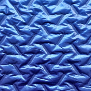Op de afbeelding: Close-up van een levendige blauwe gewatteerde stof. Het materiaal heeft een herhalend geometrisch patroon van zigzags met een getextureerde, gestippelde lijn die door elke zigzag loopt. De stof heeft een glanzende, satijnachtige uitstraling.