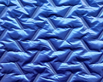 Puede incluir: Primer plano de una vibrante tela acolchada azul. El material tiene un patrón geométrico repetitivo de zigzags con una línea punteada texturizada que atraviesa cada zigzag. La tela tiene un aspecto brillante, similar al satén.