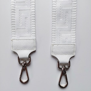 Könnte beinhalten: Zwei weiße Stoffbänder mit geometrischem Muster und weißen Lederenden. Jedes Band hat einen silbernen Drehhaken.