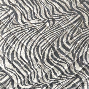 Op de afbeelding: Een gewatteerde stof met een zebra streepmotief in grijstinten en gebroken wit. De stof is gestikt in een ruitpatroon, waardoor een gestructureerd oppervlak ontstaat. Het ontwerp is een gedurfde, grafische print.