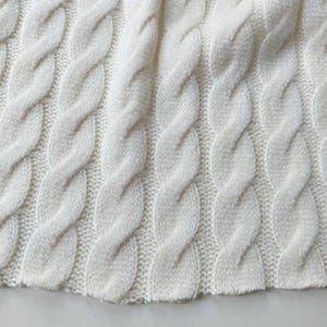 Könnte beinhalten: Nahaufnahme eines cremefarbenen Strickstoffs mit Zopfmuster. Die Textur ist weich und das Design weist vertikale, ineinander verwobene Stränge auf, die eine erhabene, strukturierte Oberfläche erzeugen. Der untere Rand ist leicht gebogen.