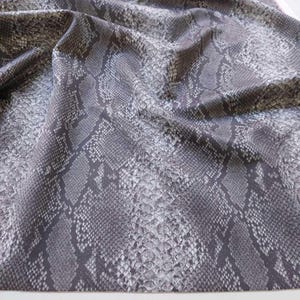 Könnte beinhalten: Nahaufnahme eines Stoffes mit Schlangenmuster. Der Stoff ist eine Mischung aus Grau und Weiß, mit einem detaillierten Druck, der die Schuppen einer Schlange nachahmt. Der Stoff ist drapiert und zeigt Falten und Textur.