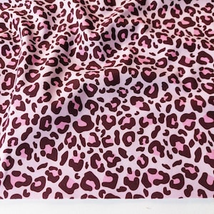 Op de afbeelding: Roze stof met een luipaardprint. De stof heeft donkerbruine en roze vlekken op een lichtroze achtergrond. De stof wordt waarschijnlijk gebruikt voor kleding of woondecoratie.