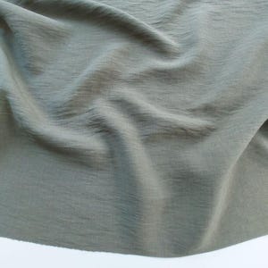 Può includere: Primo piano di un tessuto drappeggiato in un colore verde oliva tenue. Il tessuto ha un aspetto strutturato e increspato, con pieghe e ombre morbide. Il materiale appare leggero e fluido, adatto all'abbigliamento o all'arredamento.