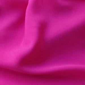 Può includere: Un primo piano di un tessuto rosa vibrante con una texture liscia e setosa. Il tessuto è drappeggiato e piegato, creando linee morbide e fluide.