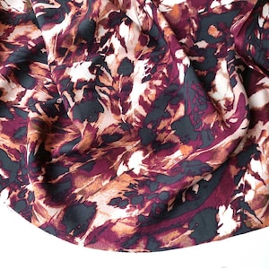 Op de afbeelding: Close-up van een stof met een abstract patroon. Het ontwerp bevat een mix van kleuren, waaronder diep bordeauxrood, donkerblauwgroen en tinten bruin en wit. De stof lijkt zacht en gedrapeerd, met zichtbare plooien en kreukels.