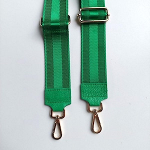 Può includere: Due cinturini in tessuto verde a righe con accessori e ganci in metallo dorato. I cinturini sono realizzati in tessuto intrecciato con un motivo a righe verde e bianco. I cinturini sono fissati a ganci in metallo dorato.