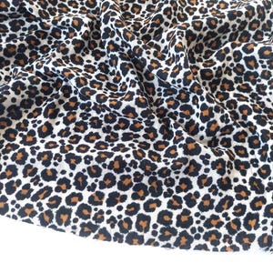 Könnte beinhalten: Nahaufnahme eines Stoffes mit Leopardenmuster. Der Stoff hat einen weißen Hintergrund mit schwarzen und braunen Flecken. Der Stoff ist gefaltet und drapiert, wodurch die Textur und das Muster sichtbar werden.