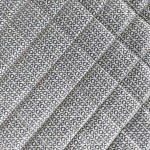 Op de afbeelding: Close-up van een gewatteerde stof met een herhalend geometrisch patroon. Het patroon heeft kleine, donkergrijze vormen op een lichtgrijze achtergrond. Diagonale stiksels creëren een rasterachtig effect over de stof.