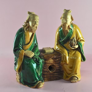 May include: Two ceramic figurines of seated Asian figures. One figure wears a green robe, the other a yellow robe. They sit on a brown log with a book. The figures have long beards and are in a contemplative pose.