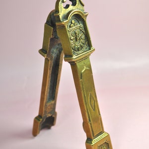 May include: A brass nutcracker shaped like a tall grandfather clock. The front features a detailed clock face with Roman numerals and a small diamond-shaped accent on the lower section. The base is decorated with a small fleur-de-lis emblem.