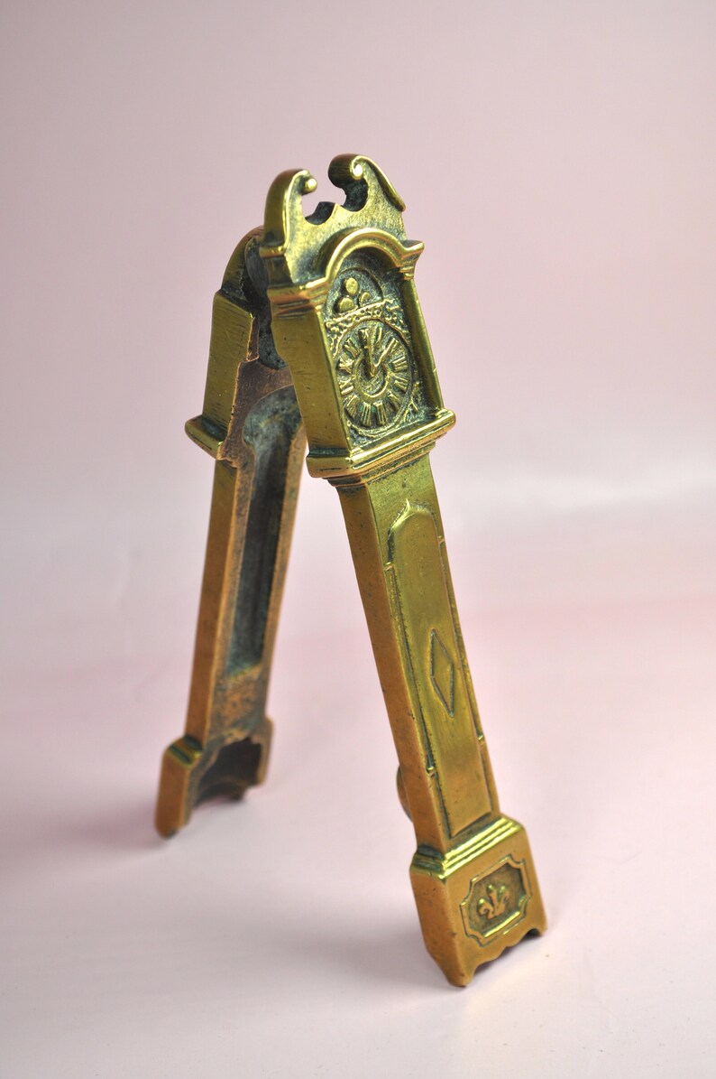 May include: A brass nutcracker shaped like a tall grandfather clock. The front features a detailed clock face with Roman numerals and a small diamond-shaped accent on the lower section. The base is decorated with a small fleur-de-lis emblem.