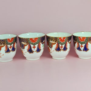 May include: Four white teacups with intricate patterns. The cups feature a design with red, blue, and green accents, and gold trim. The teacups are arranged in a row against a pink background, showcasing their detailed craftsmanship.