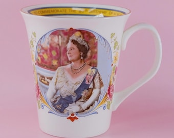 May include: A white ceramic mug featuring a portrait of Queen Elizabeth II in a decorative oval frame. The rim text reads TO COMMEMORATE THE 80TH BIRTHDAY OF H.M. QUEEN ELIZABETH II. The design includes floral accents and a blue sash on the portrait.