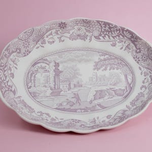 May include: An oval ceramic platter with a scalloped edge, featuring a white base and a detailed purple transferware print. The central scene depicts a landscape with ruins and figures, surrounded by a wide border of floral and scroll patterns.