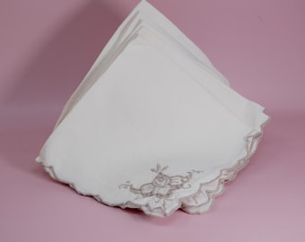 May include: A stack of white square cloth napkins featuring scalloped edges and light brown floral embroidery in one corner. The napkins are folded into a triangular shape, showcasing the detailed stitching and the clean white fabric texture.