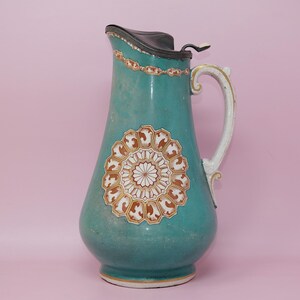 May include: A teal ceramic pitcher featuring a dark metal hinged lid, a curved handle, and a central circular medallion with white and orange geometric patterns. A decorative band with similar orange and white motifs encircles the upper neck.