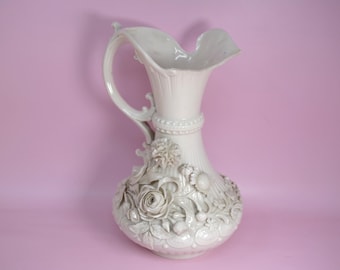 May include: A white ceramic pitcher featuring a flared, ruffled rim and a curved handle. The body is decorated with intricate, raised floral relief patterns, including roses and leaves, and vertical fluting on the neck and base.