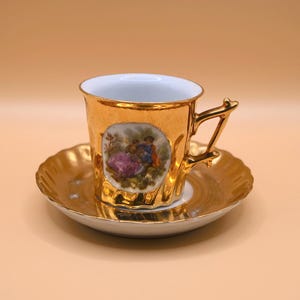 Puede incluir: Un juego de taza y platillo dorados. La taza presenta un asa decorativa y una escena pintada de una pareja. El platillo y la taza tienen un borde festoneado. El juego es de porcelana.