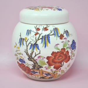 May include: A round white ceramic jar with a matching lid, featuring a colorful hand-painted design of red peonies, green leaves, and blue and yellow hanging floral clusters, set against a white background with a small wooden fence detail.